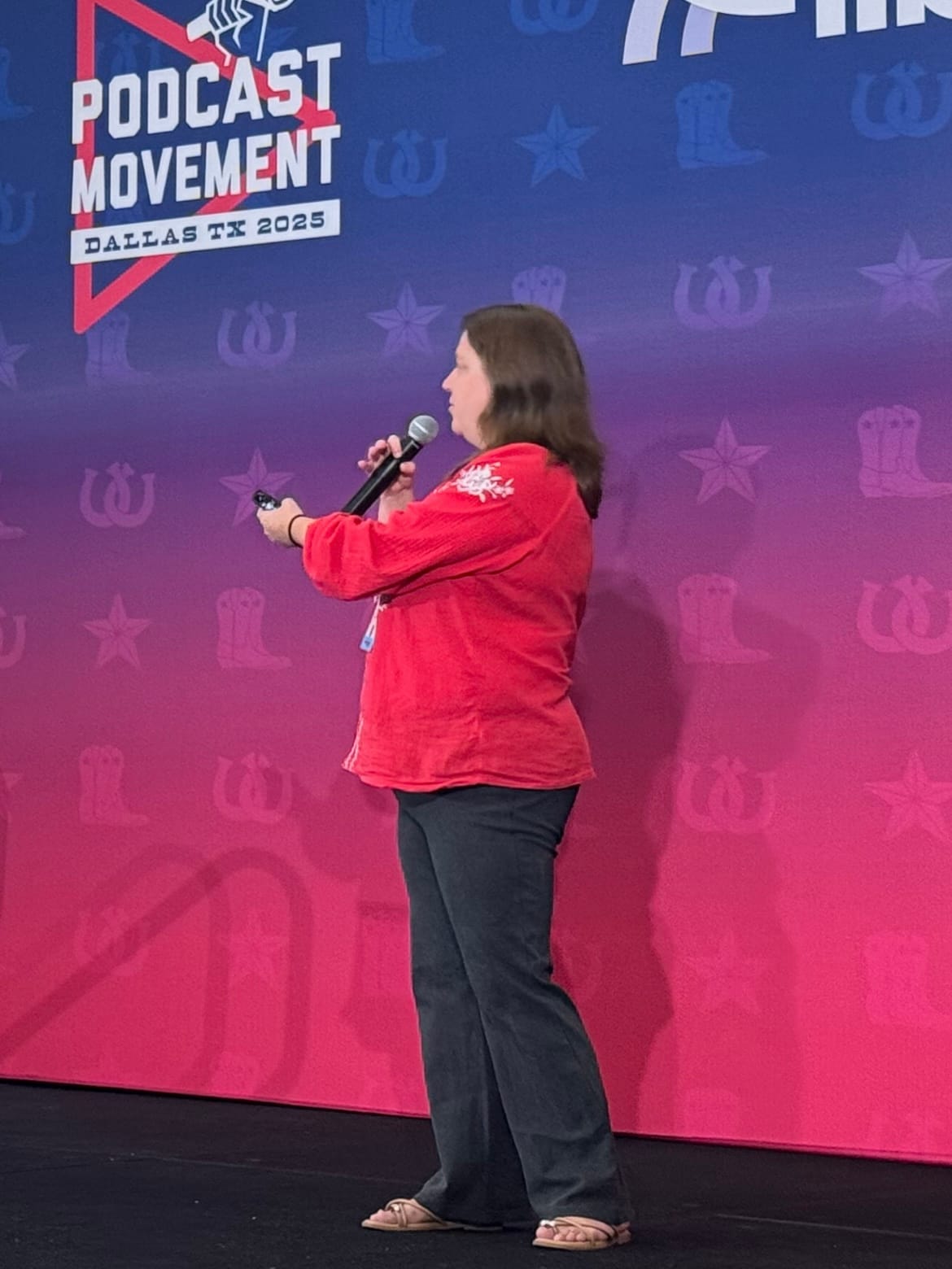 A person in a red shirt stands on stage at a conference, speaking into a microphone. The backdrop reads "Podcast Movement Dallas TX 2025."