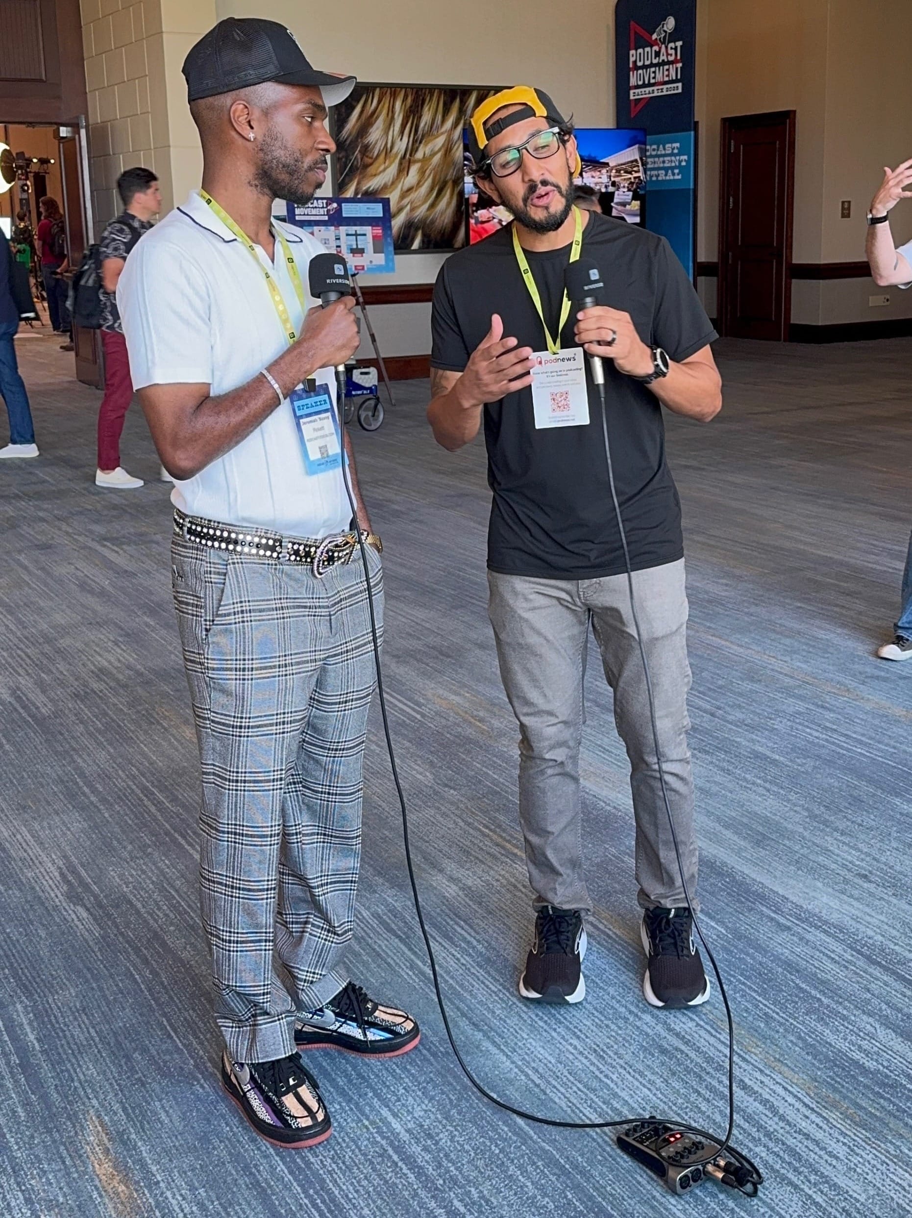 Two people are engaged in an interview at a conference. One holds a microphone, while both wear badges. A "Podcast Movement" sign is visible in the background.