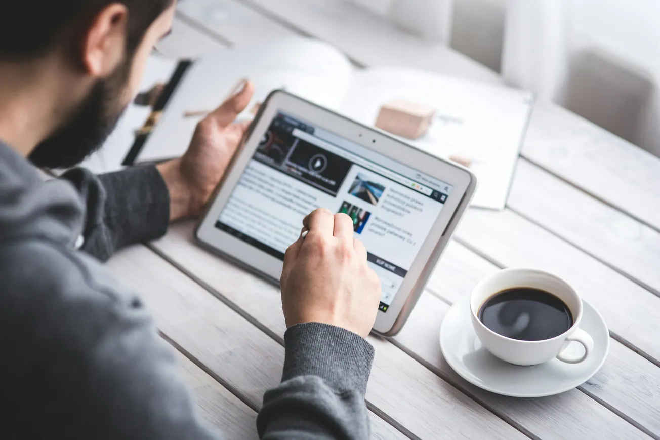 A man scrolling through a news website on his iPad with a cup of coffee nearby and a book next to him on the wooden table.