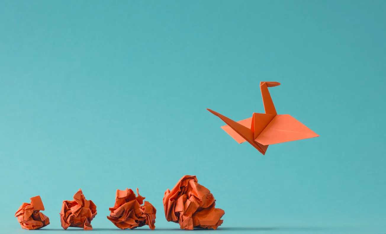 Transformation concept with orange origami crane rising above crumpled paper – Symbol of creativity, growth, and innovation.
