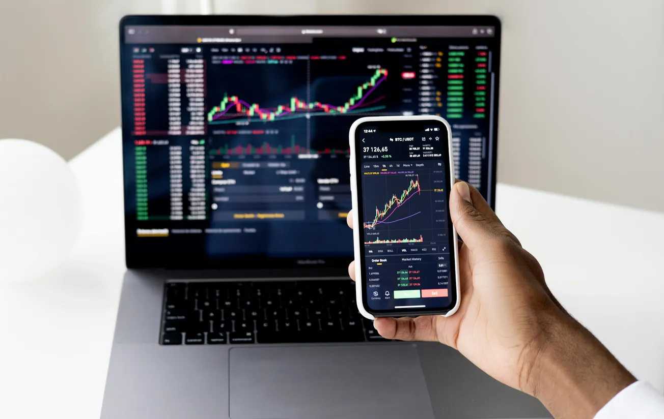 A person holds a smartphone displaying stock market charts in front of a laptop with similar charts.