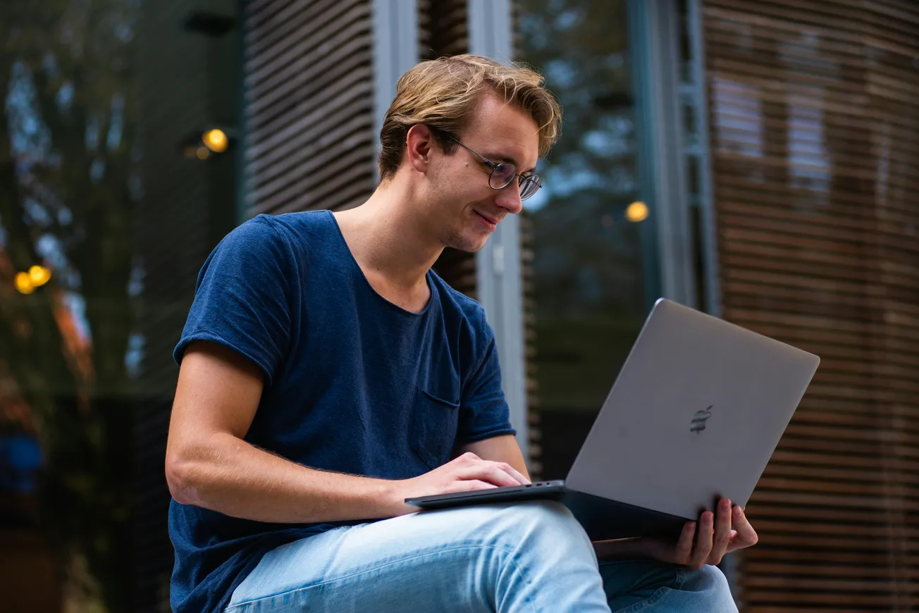 A man in glasses sits outside on stairs, engaged with a laptop. He's wearing a blue shirt and jeans.