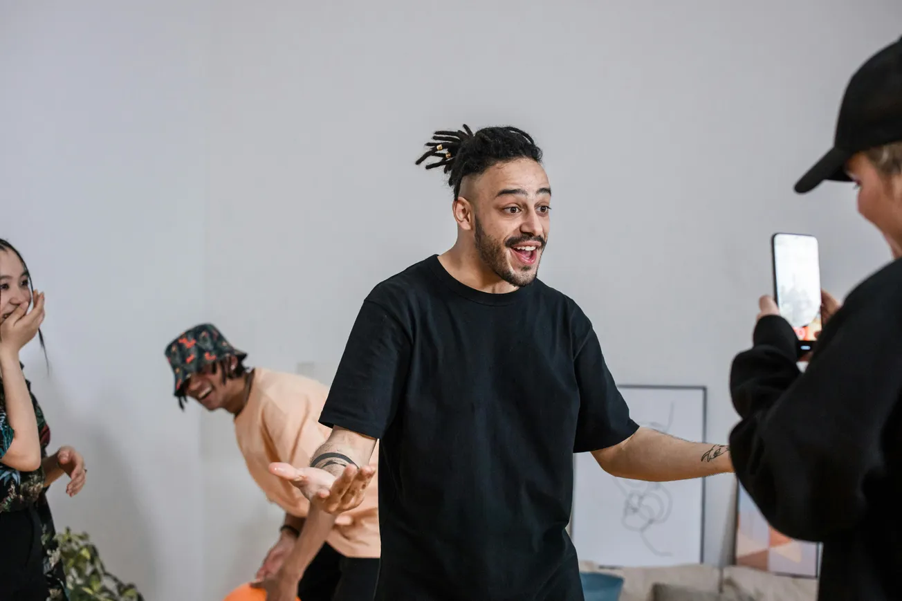 A man in a black shirt appears surprised and amused while friends laugh and record him on a phone.