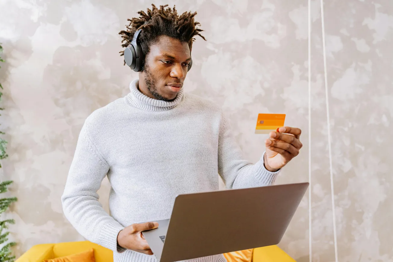 A person wearing headphones and a gray sweater holds a credit card while looking at a laptop. The setting is a cozy room with a yellow couch.