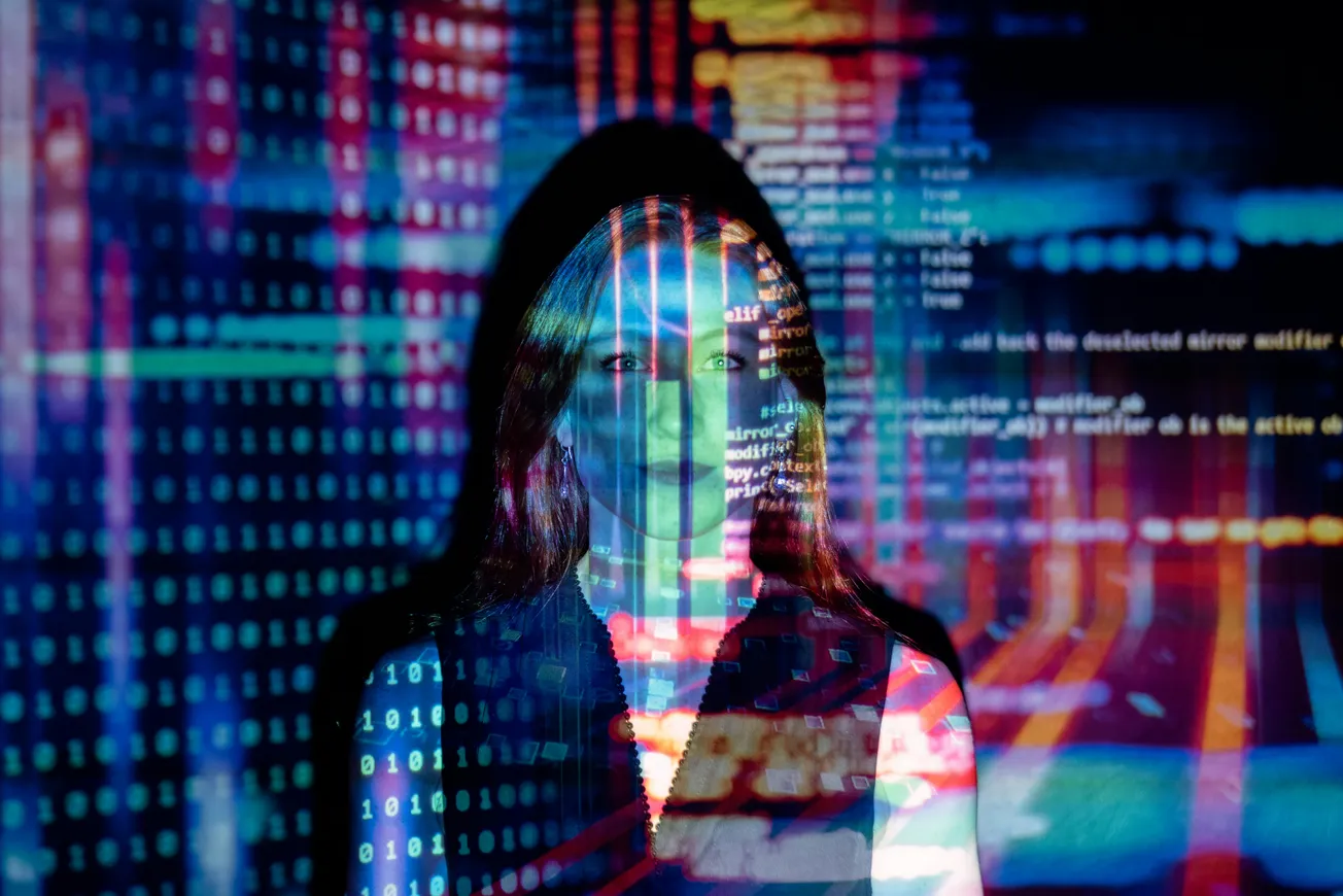 A woman stands in front of a colorful digital projection with binary code and programming language displayed across her face and body.