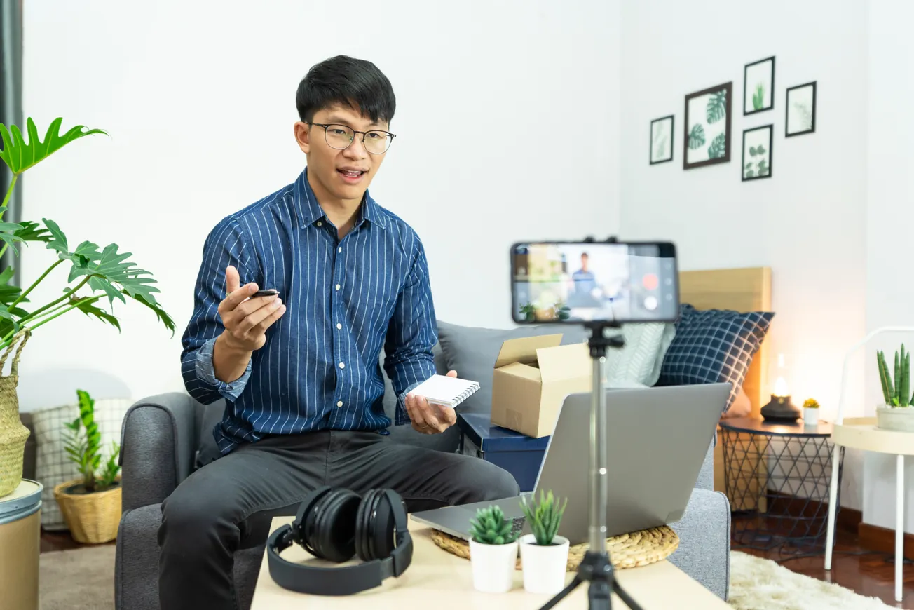 Man recording video content in a cozy room with plants. He gestures while holding a notepad. Camera, laptop, and headphones nearby convey a tech-savvy atmosphere.