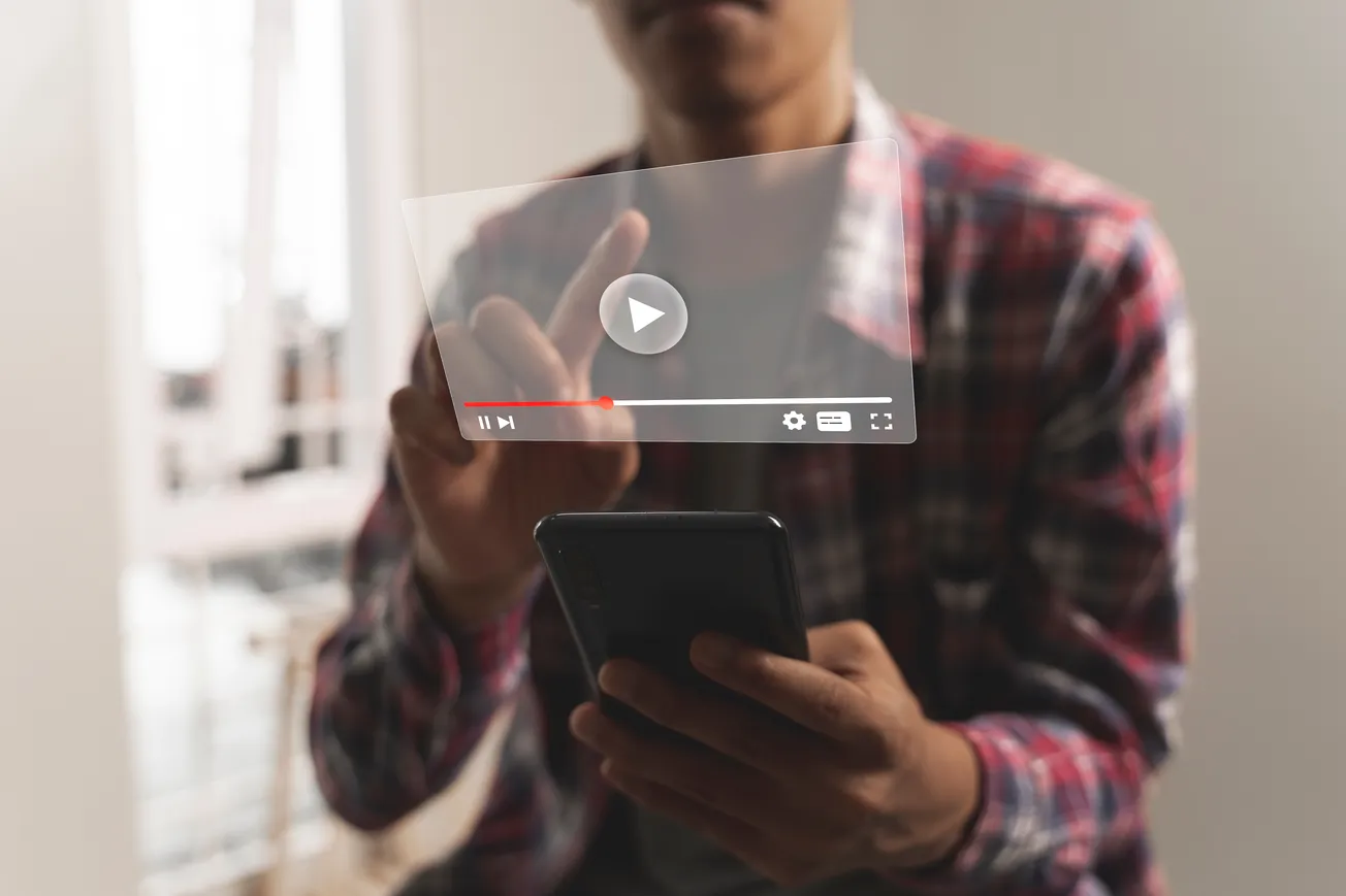 A person wearing a plaid shirt interacts with a holographic video play interface while holding a smartphone.