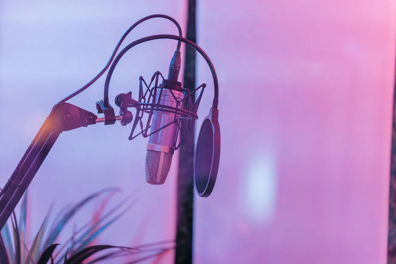 A studio microphone with a pop filter is suspended on an adjustable arm against a softly lit pink and purple background, conveying a creative atmosphere.