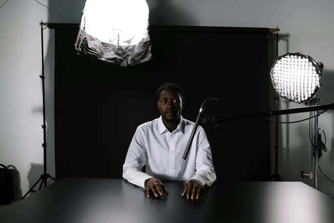 A person in a white shirt sits at a table in a dimly lit room, surrounded by professional studio lighting and a microphone.