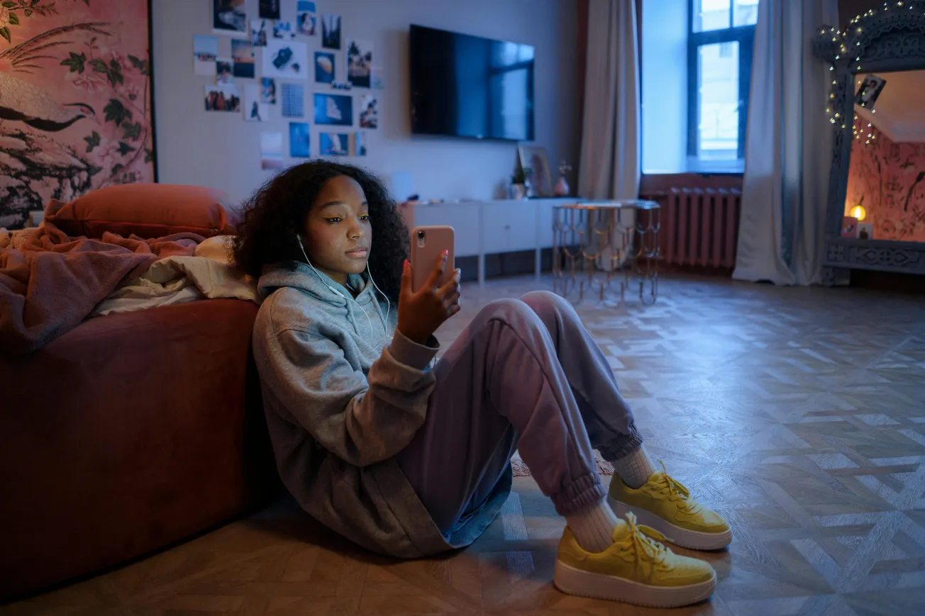 A young person in a gray hoodie and yellow sneakers sits on the floor, focused on a smartphone. The cozy room has warm lighting, a large TV, and photos on the wall.