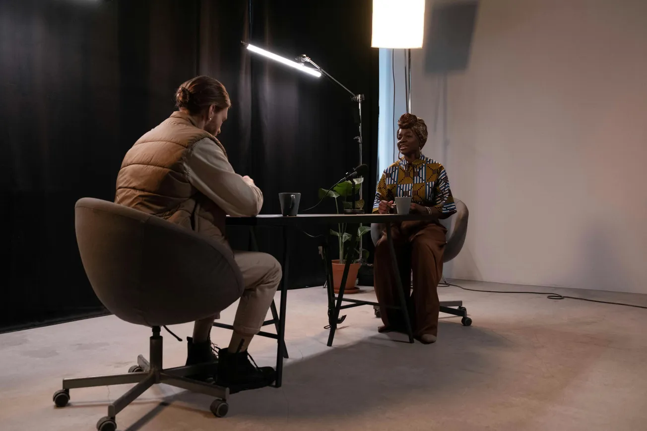 Two people sit across a table in a dimly lit room, engaged in conversation. One person wears a patterned shirt, and a potted plant sits between them.