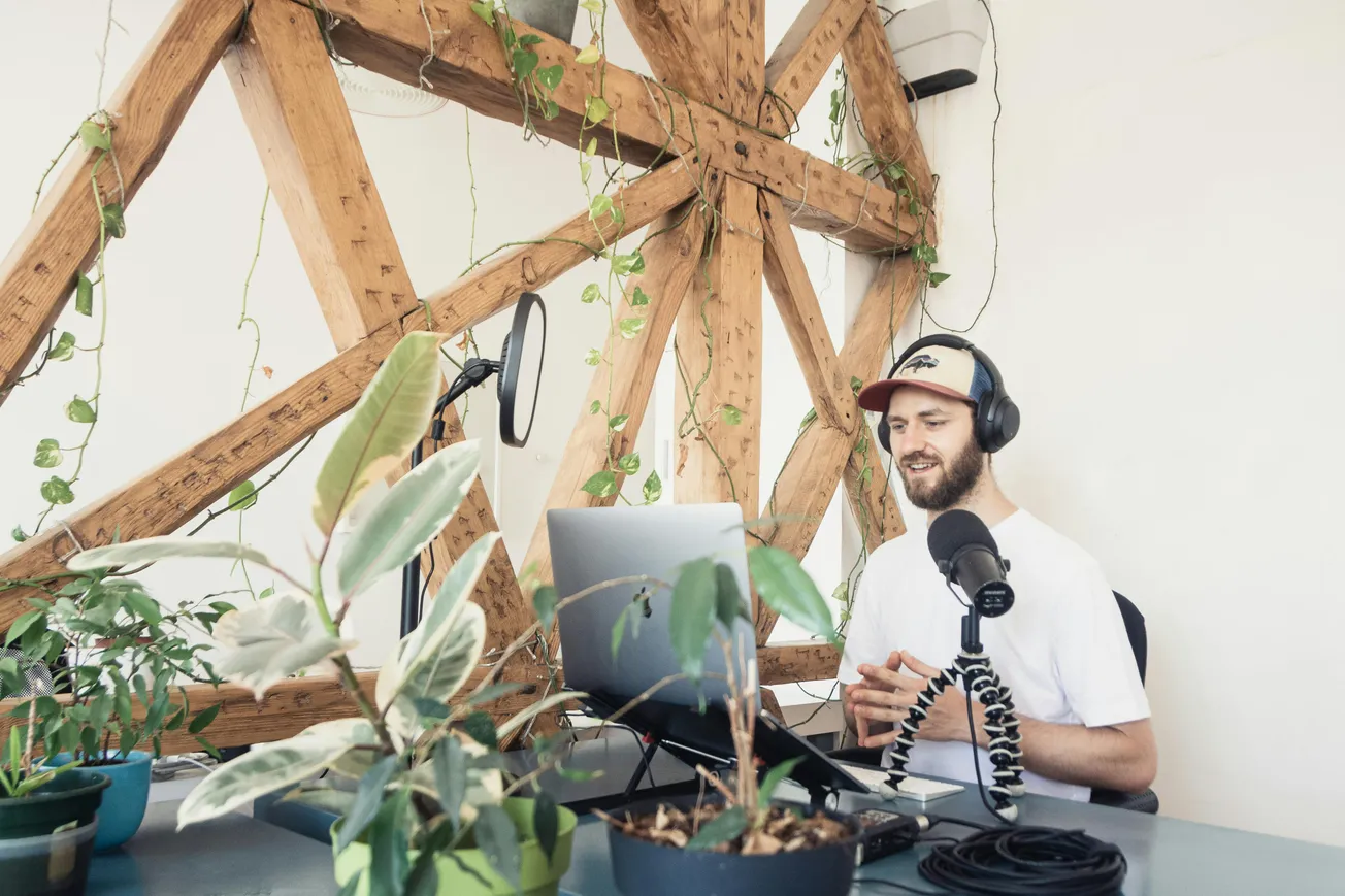 Man with beard wearing headphones and cap recording podcast at a desk. He is in a plant-filled studio with rustic wooden beams and a microphone.