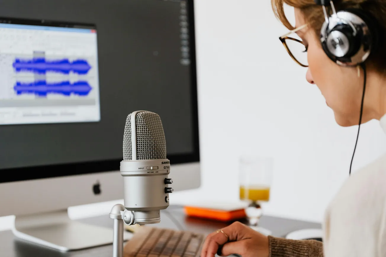 A person wearing headphones works at a computer with audio editing software on the screen. A microphone is prominently placed in the foreground.
