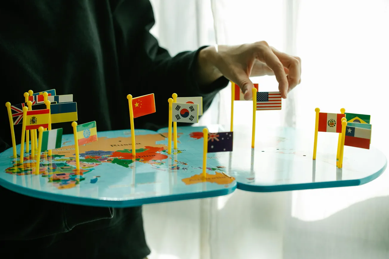 A person holding a world map board with small flags representing different countries. The hand gently adjusts the U.S. flag.