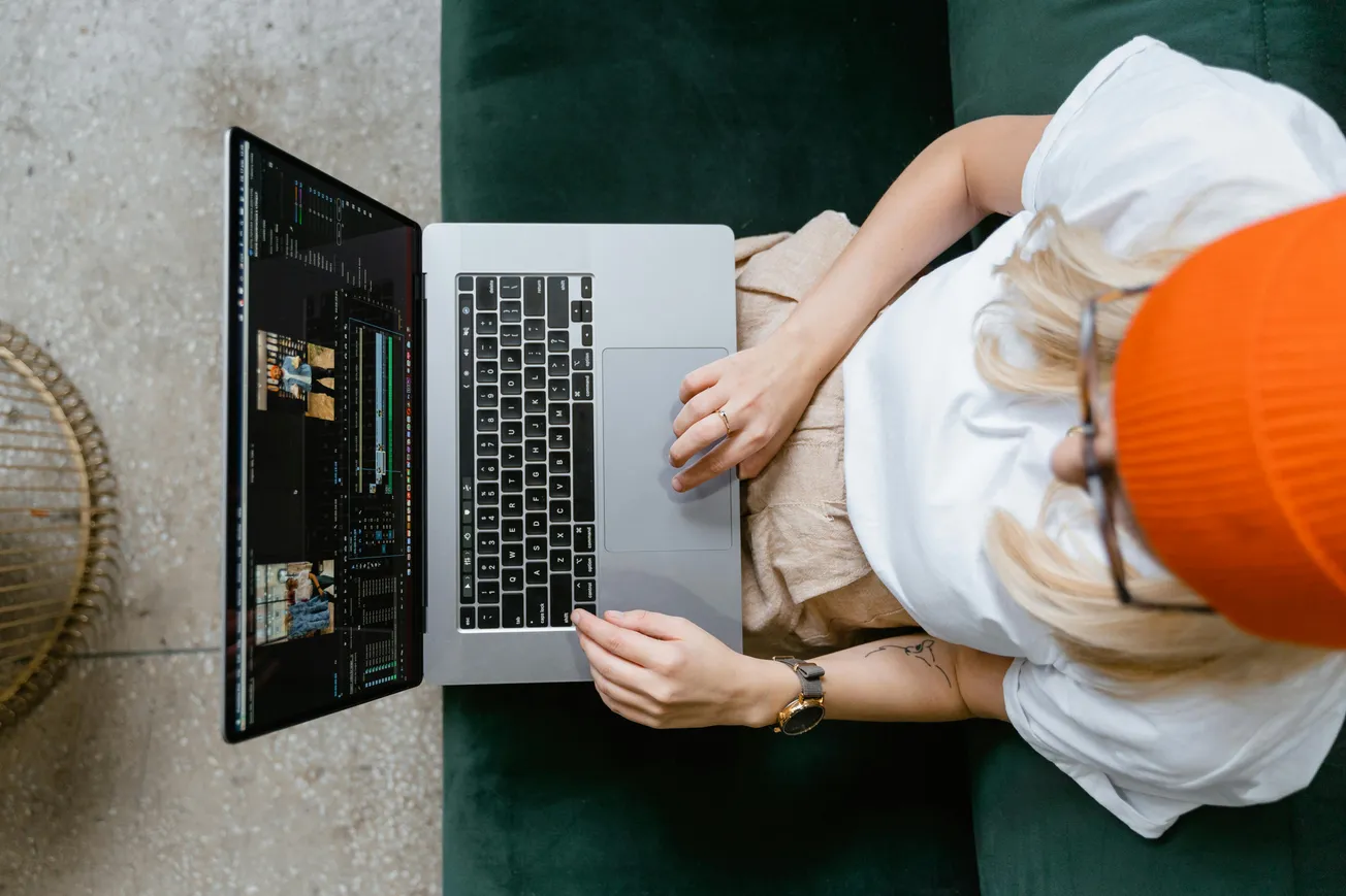 Overhead view of a person in an orange beanie editing video on a laptop. They sit on a dark green couch, conveying focus and creativity.