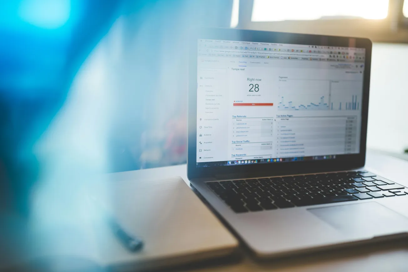 Laptop displaying analytics software with graphs and user data. The screen shows "Right now: 28" users. A notebook is partially visible nearby.