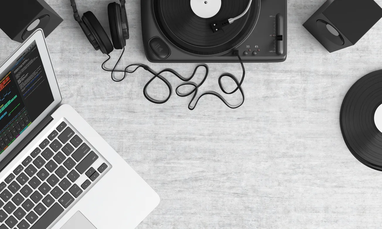 Aerial view of a turntable with a spinning vinyl record, headphones, speakers, and a laptop with code on the screen, set on a light gray surface.