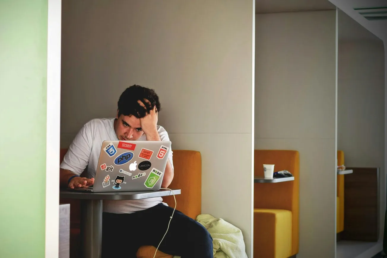 A person sits in a booth, looking frustrated, with one hand on their head while working on a laptop covered in tech stickers, embodying stress.