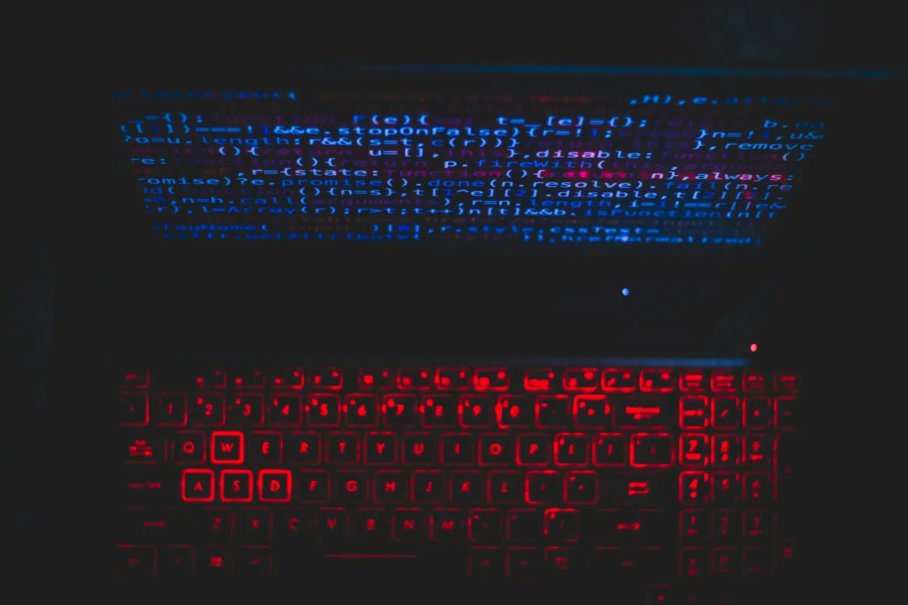 A laptop with red backlit keys shows blue code on its screen in a dark room, evoking a mysterious and high-tech atmosphere.
