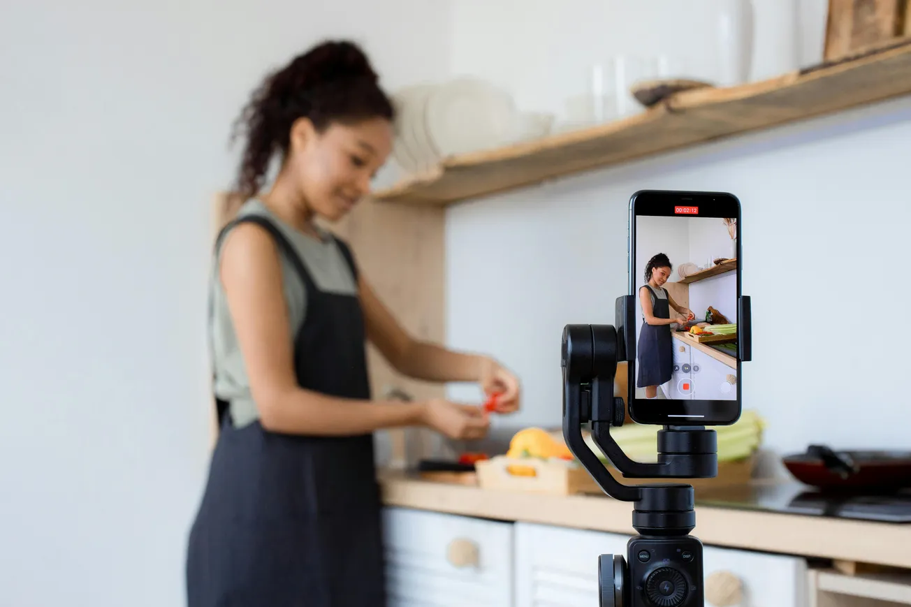 A woman in a kitchen chops vegetables on a counter. She's being filmed by a smartphone on a tripod, capturing her cooking activity.