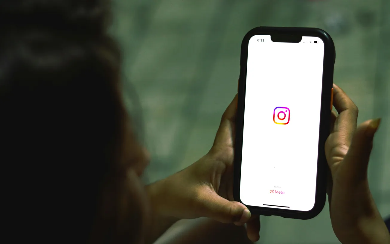 A person holding a smartphone showing the Instagram logo on the screen. The background is blurred, creating a focused view on the phone's display.