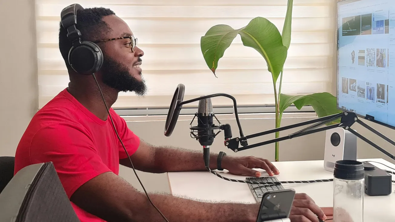 A person in a red shirt, wearing headphones, sits at a desk with a microphone, speaking into it while using a computer. A large plant is in the background.