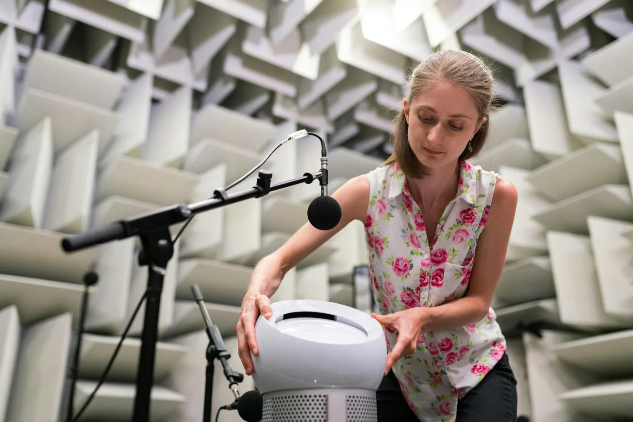 A woman in a floral shirt adjusts a device in an anechoic chamber, surrounded by sound-absorbing panels. A microphone is positioned nearby.