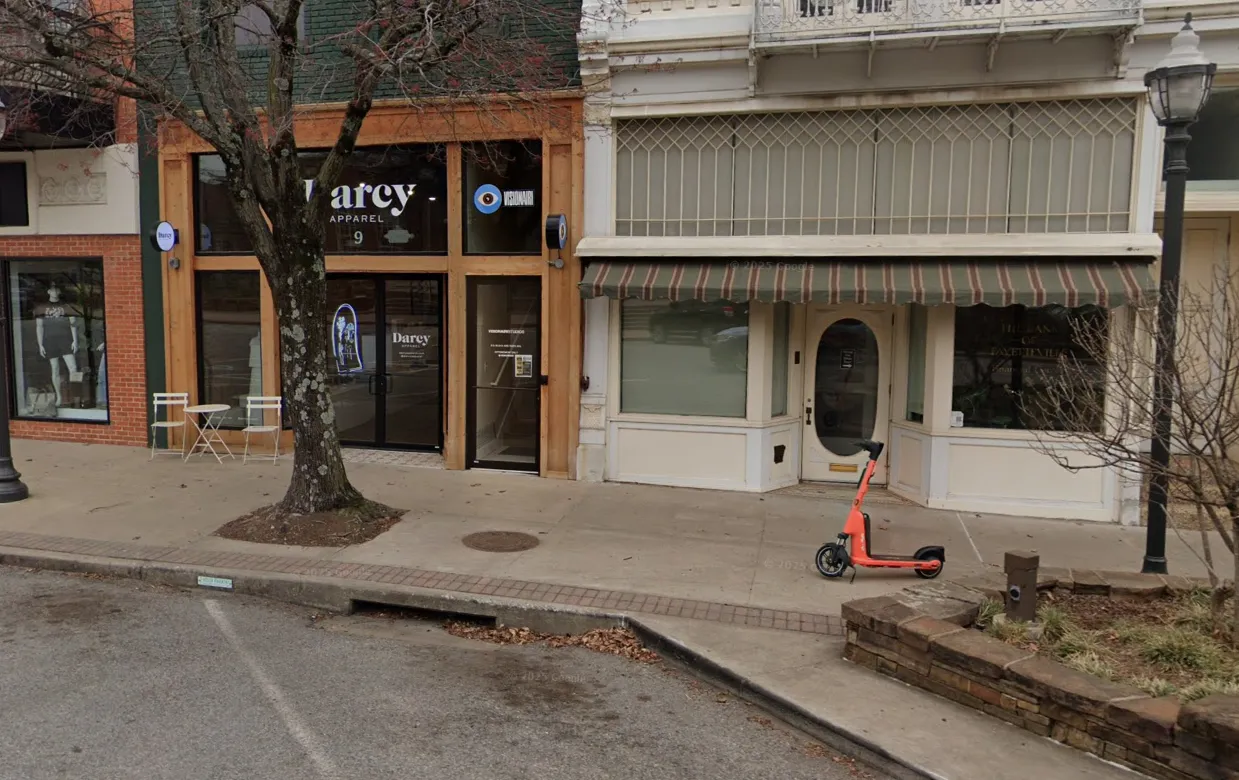 Storefronts on a quiet street include a clothing shop with a large window display and a closed store with a striped awning. An orange scooter is parked nearby.
