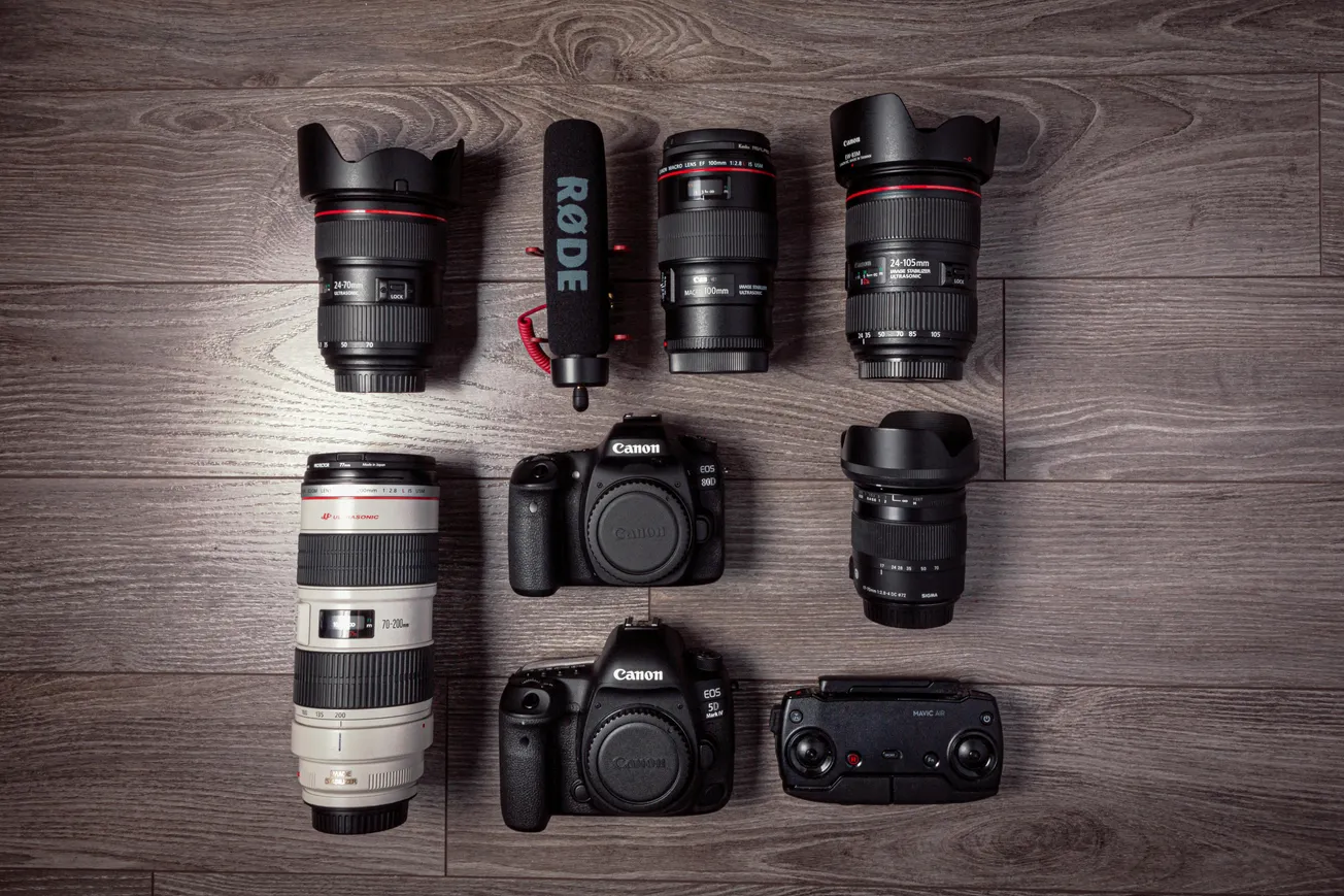 A collection of photography gear on a wooden floor, including six camera lenses, two Canon DSLRs, a Rode microphone, and a drone controller.