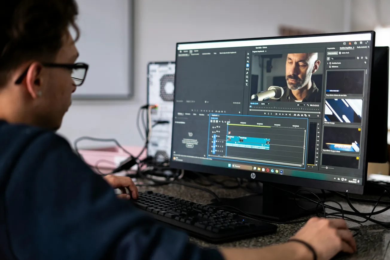 A person wearing glasses edits a video on a large computer monitor, showing a man speaking into a microphone.