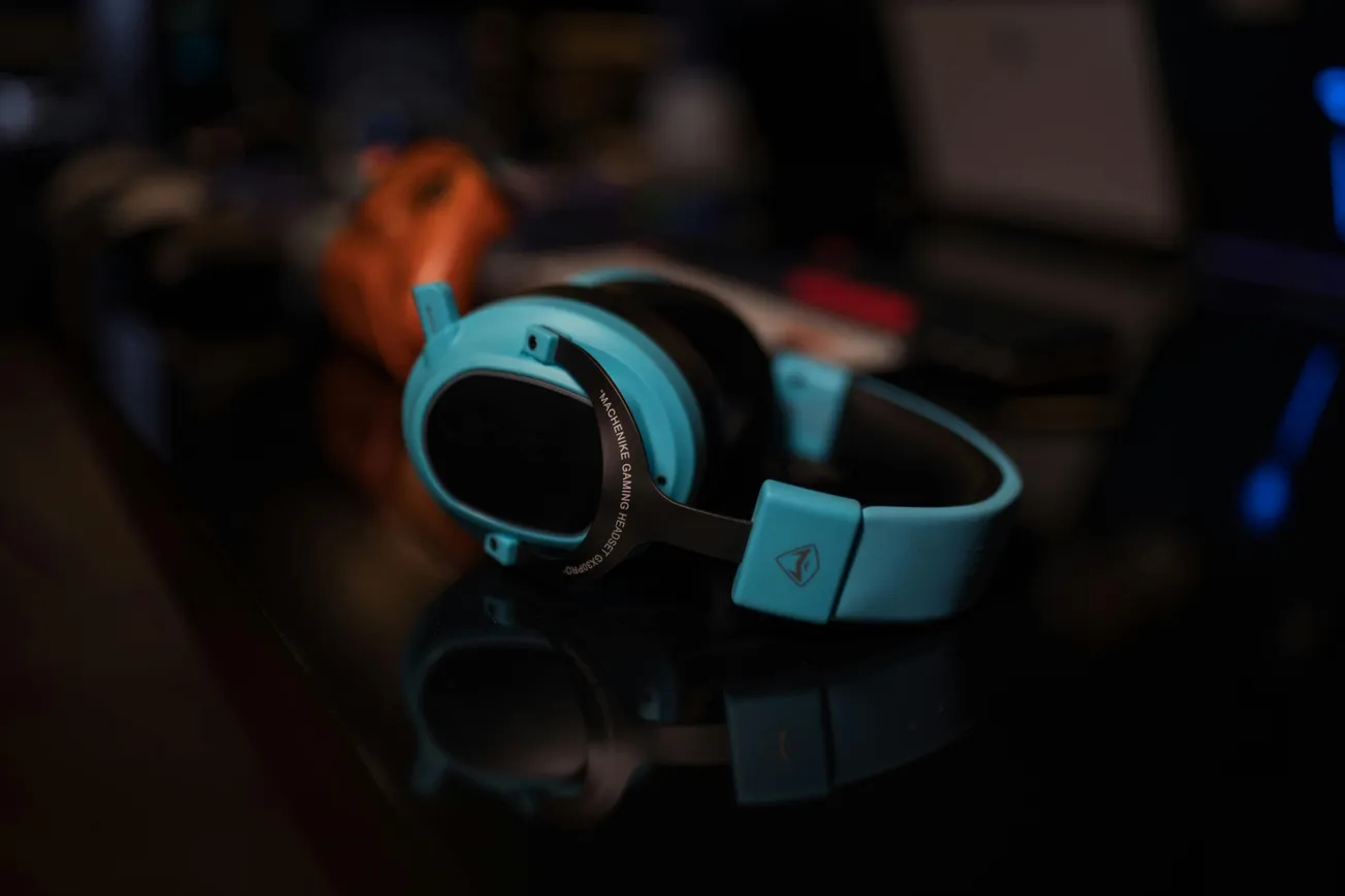 Turquoise headphones rest on a dark reflective surface, surrounded by blurred gadgets. The scene conveys a tech-focused, modern ambiance.