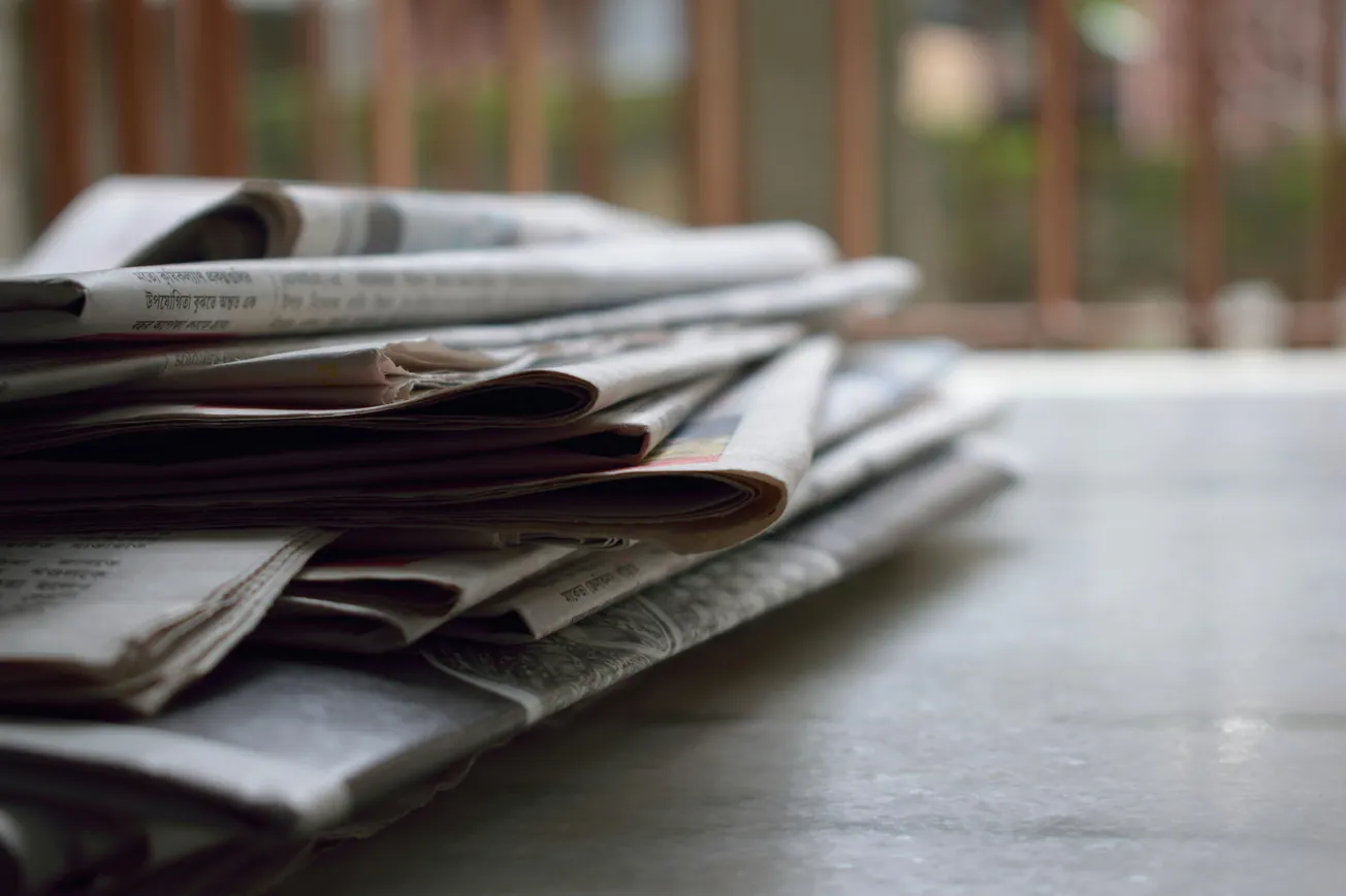A stack of newspapers lies on a polished surface, with soft sunlight filtering through window blinds in the background.