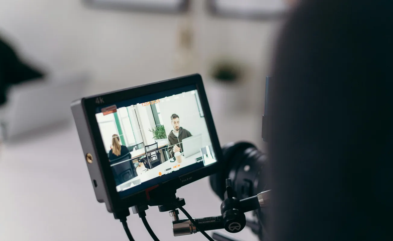 A camera monitor displays a film scene with a man and woman sitting at a desk, surrounded by paperwork. 
