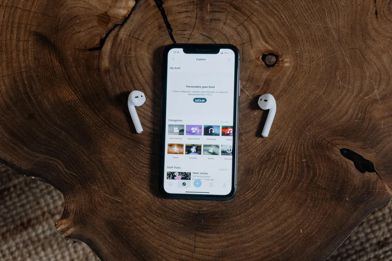 Smartphone displaying a music app on a wooden surface, flanked by two wireless earbuds. The natural wood texture adds warmth to the tech setup.