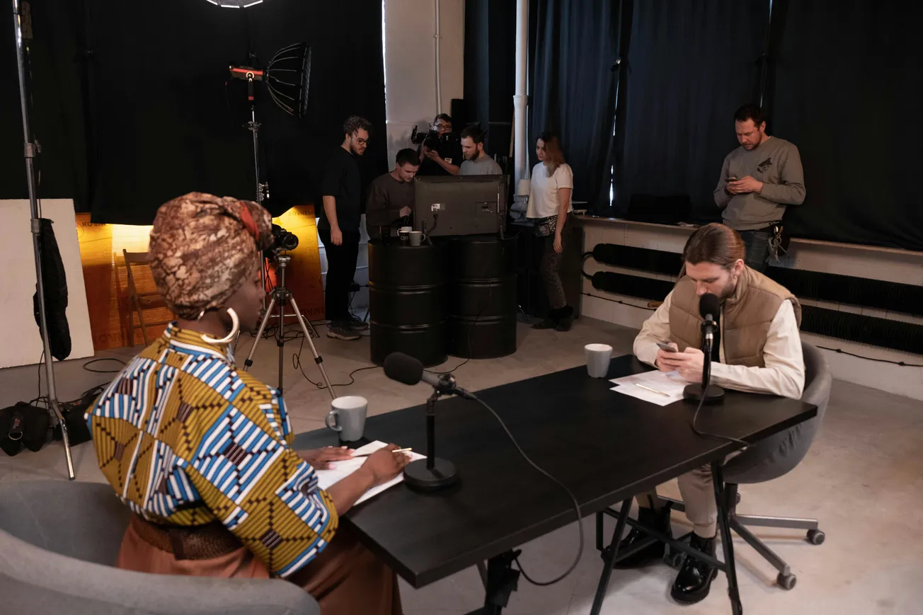 Two people sit across a table with microphones, preparing for a podcast. A group of technicians sets up equipment in the background.
