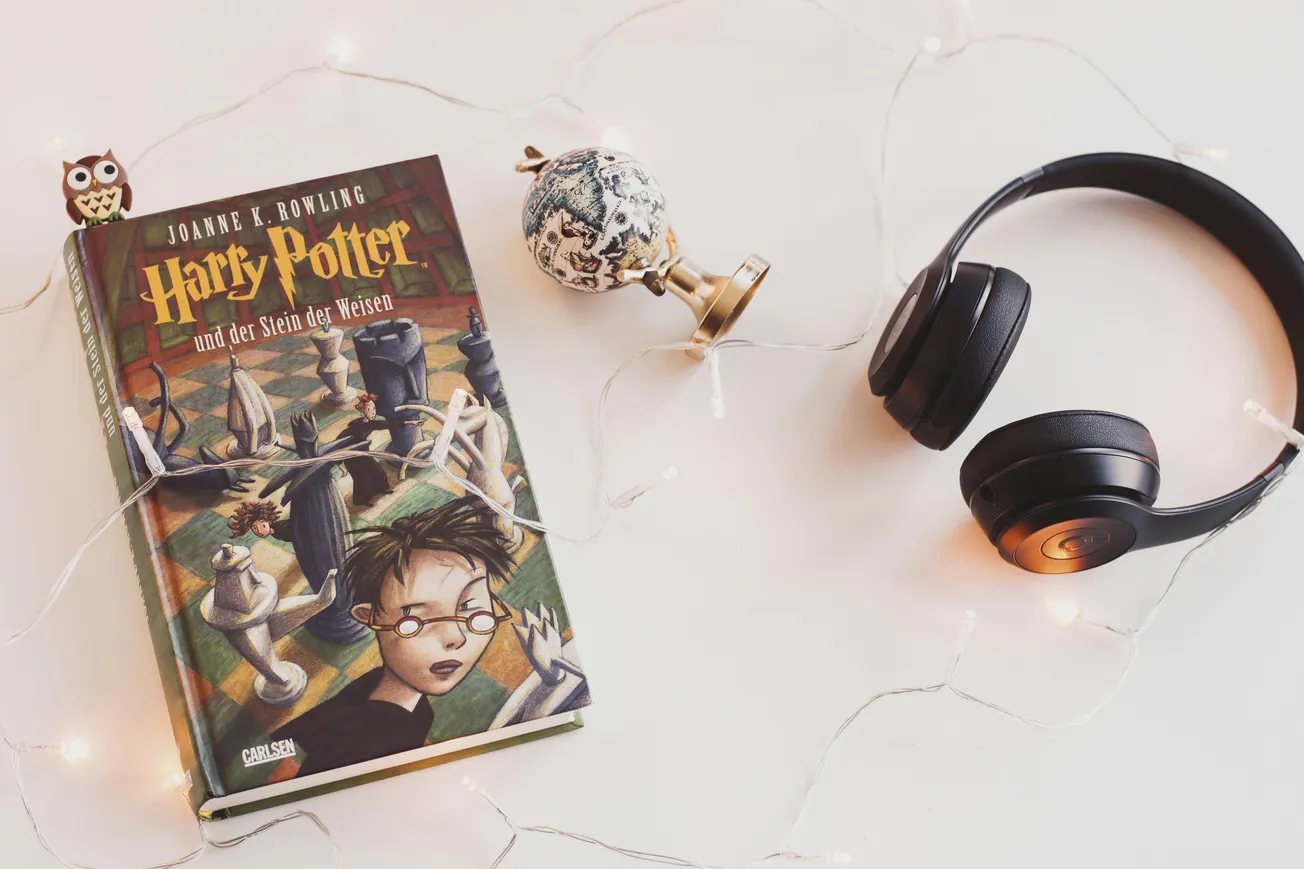 Book "Harry Potter und der Stein der Weisen" on a table with headphones, fairy lights, a globe ornament, and an owl figurine, creating a magical ambiance.