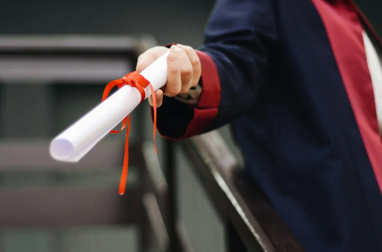 A person in a navy and red academic gown holds out a diploma tied with a red ribbon, symbolizing achievement and celebration, against a blurred background.
