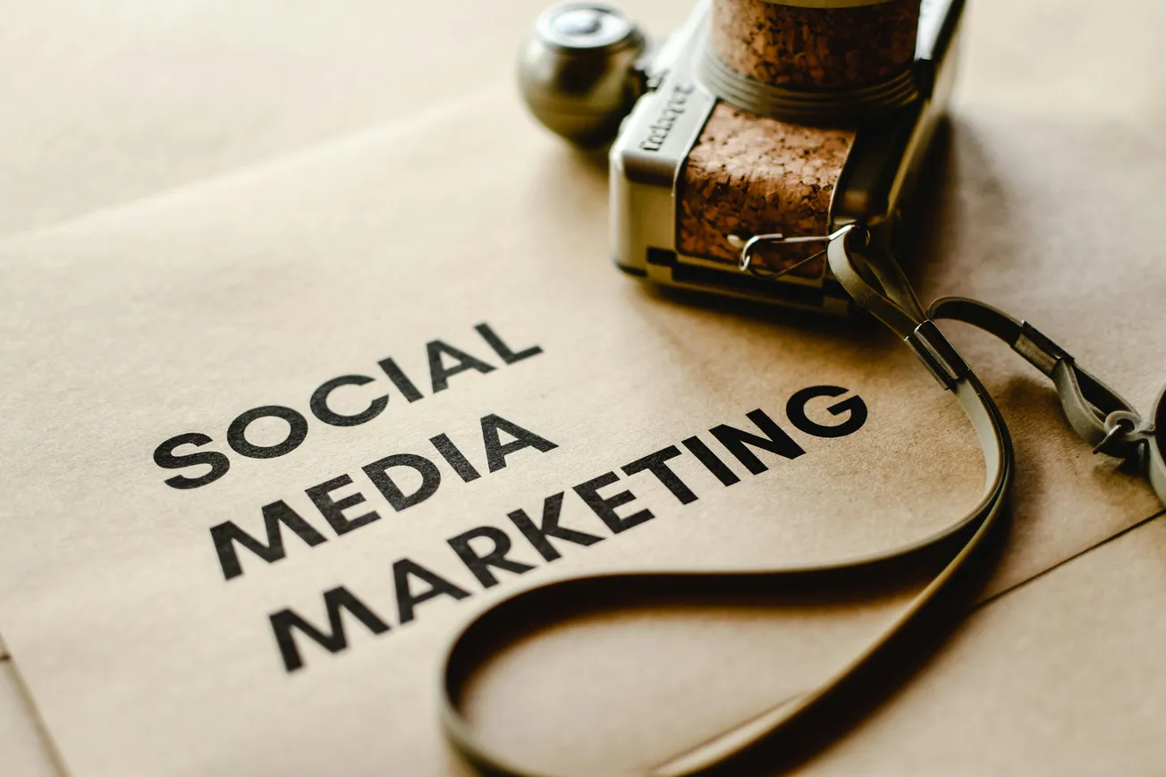 Brown paper with "Social Media Marketing" text in bold black letters. A vintage camera with a strap is positioned at the top right.