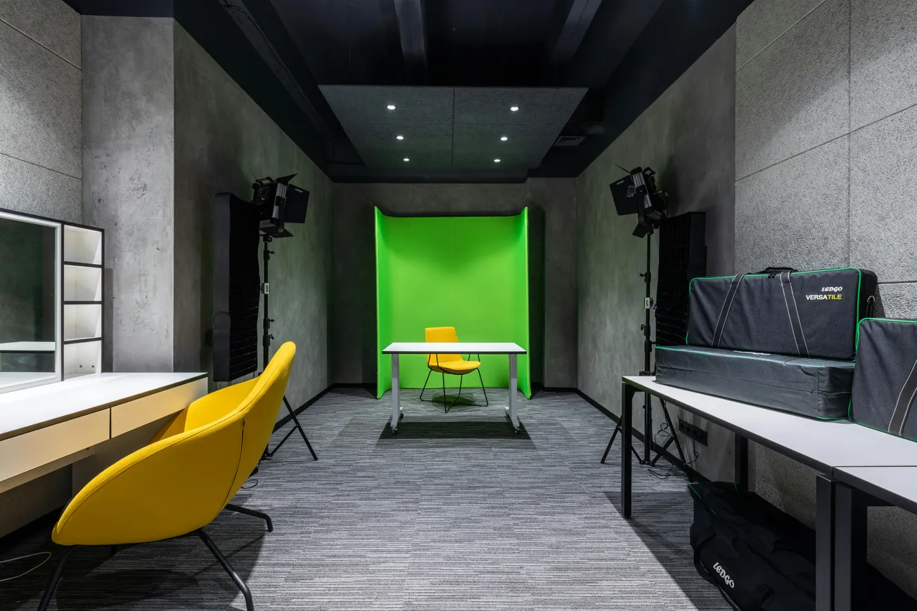 Small studio setup featuring a green screen backdrop, a table, a yellow chair, two studio lights, storage cases, and a vanity with a large mirror.