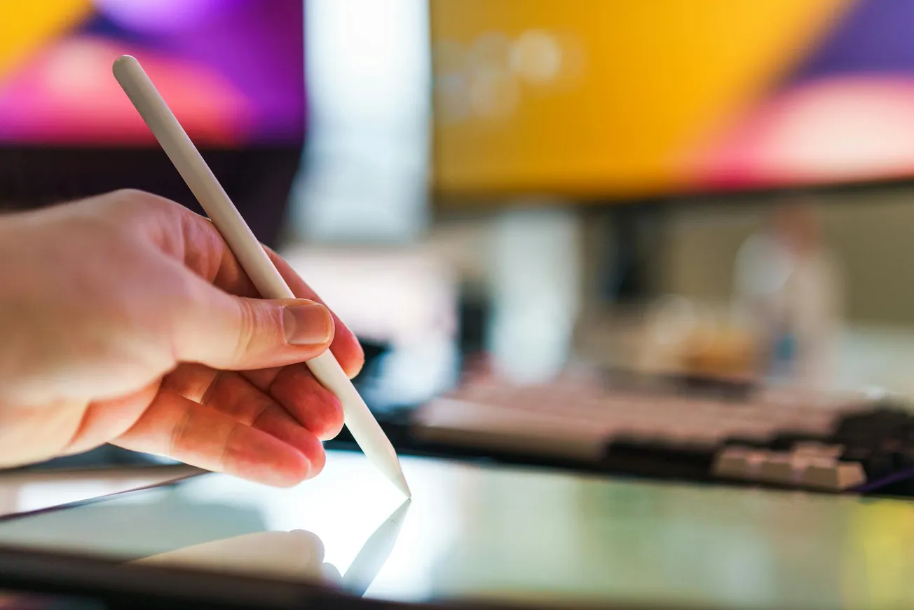 A hand uses a white stylus on a tablet screen, with vibrant monitors blurred in the background.
