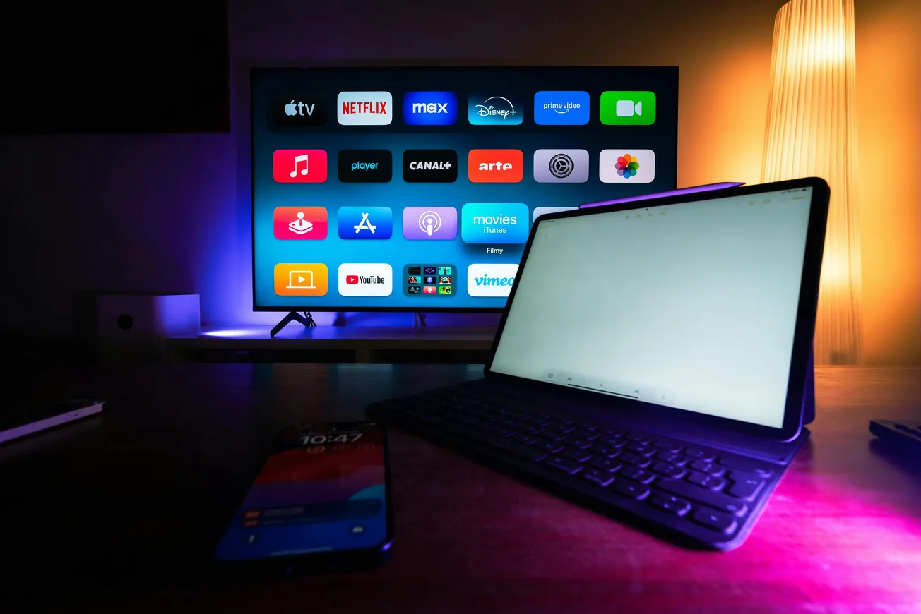Dimly lit room with a tablet on a keyboard stand in the foreground and a smartphone. A TV displays streaming app icons.