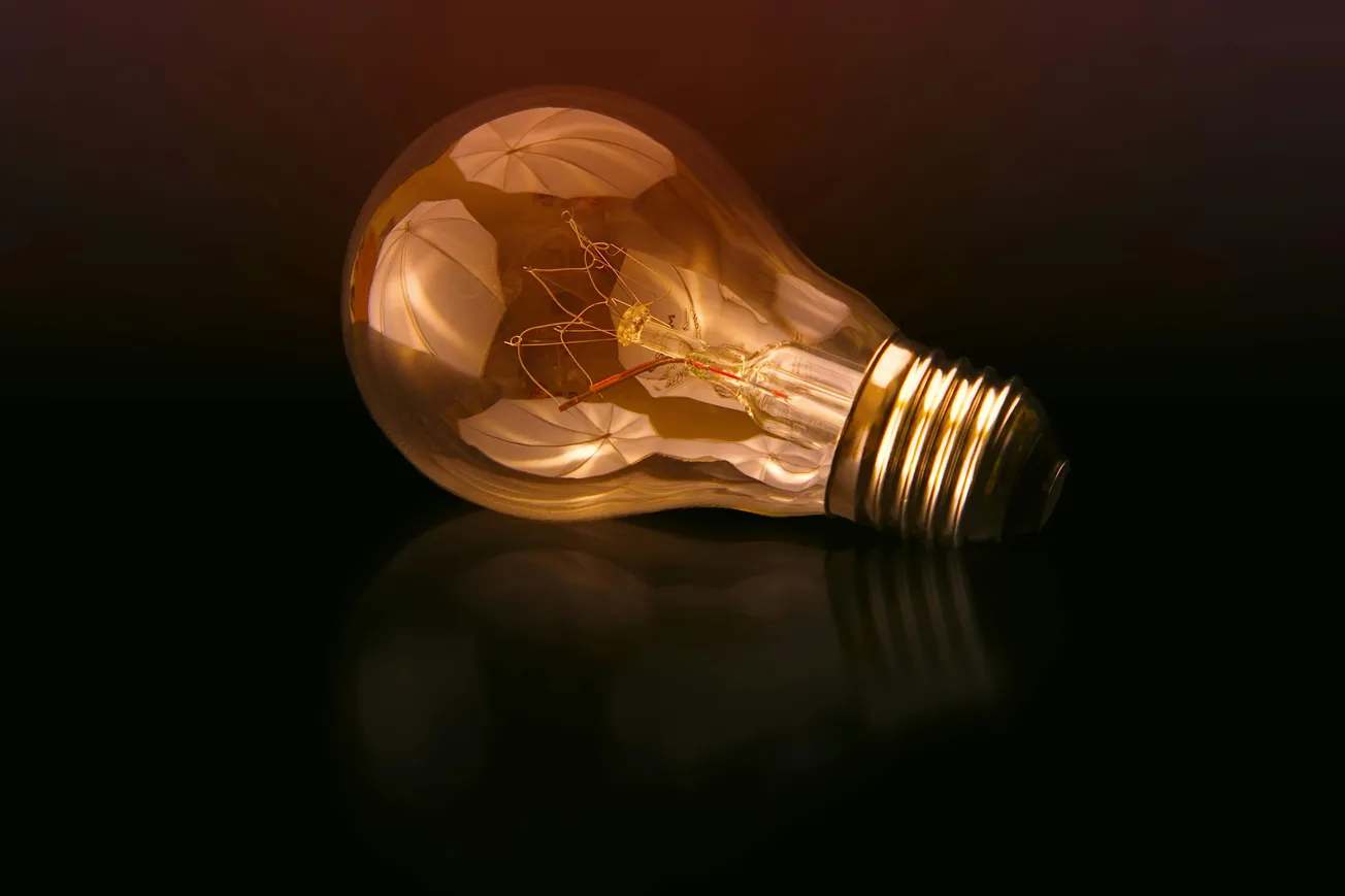 A glowing incandescent light bulb lies on a dark reflective surface, casting a warm amber hue. The mood is contemplative and innovative.