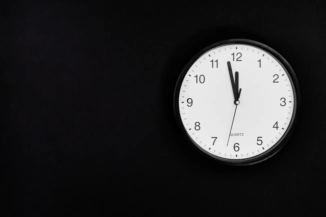 A white wall clock with black hands and numbers is set against a dark background. The minimalist design evokes a simple and modern feel.