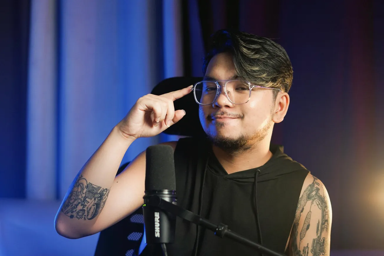 A person with glasses and tattoos smiles, pointing to temple near a microphone.