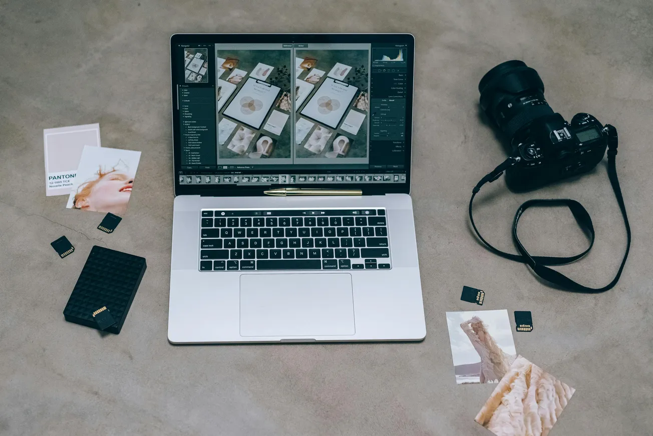 A laptop on a beige surface shows an editing software with images. Surrounding it are a DSLR camera, memory cards, a portable drive, and photo prints.