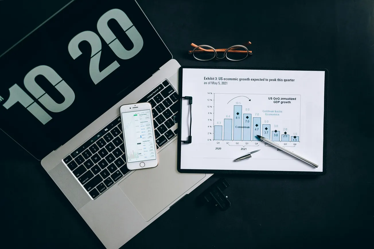 A laptop with a screen showing "10:20," a smartphone displaying data, and a clipboard with a financial graph are on a black surface. Reading glasses and a pen are nearby.