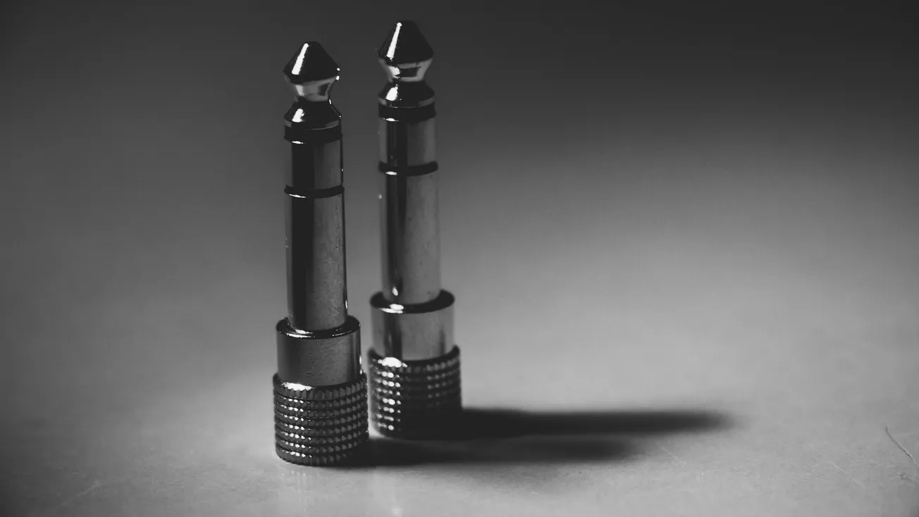 Two metallic audio jack plugs stand upright on a smooth surface, casting soft shadows. The monochrome tone gives a sleek, modern, and minimalist feel.