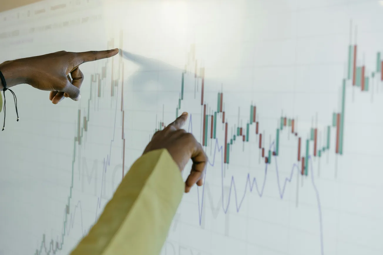 Two hands point at a financial chart on a screen, indicating key data points. The image conveys analysis and decision-making in a business context.