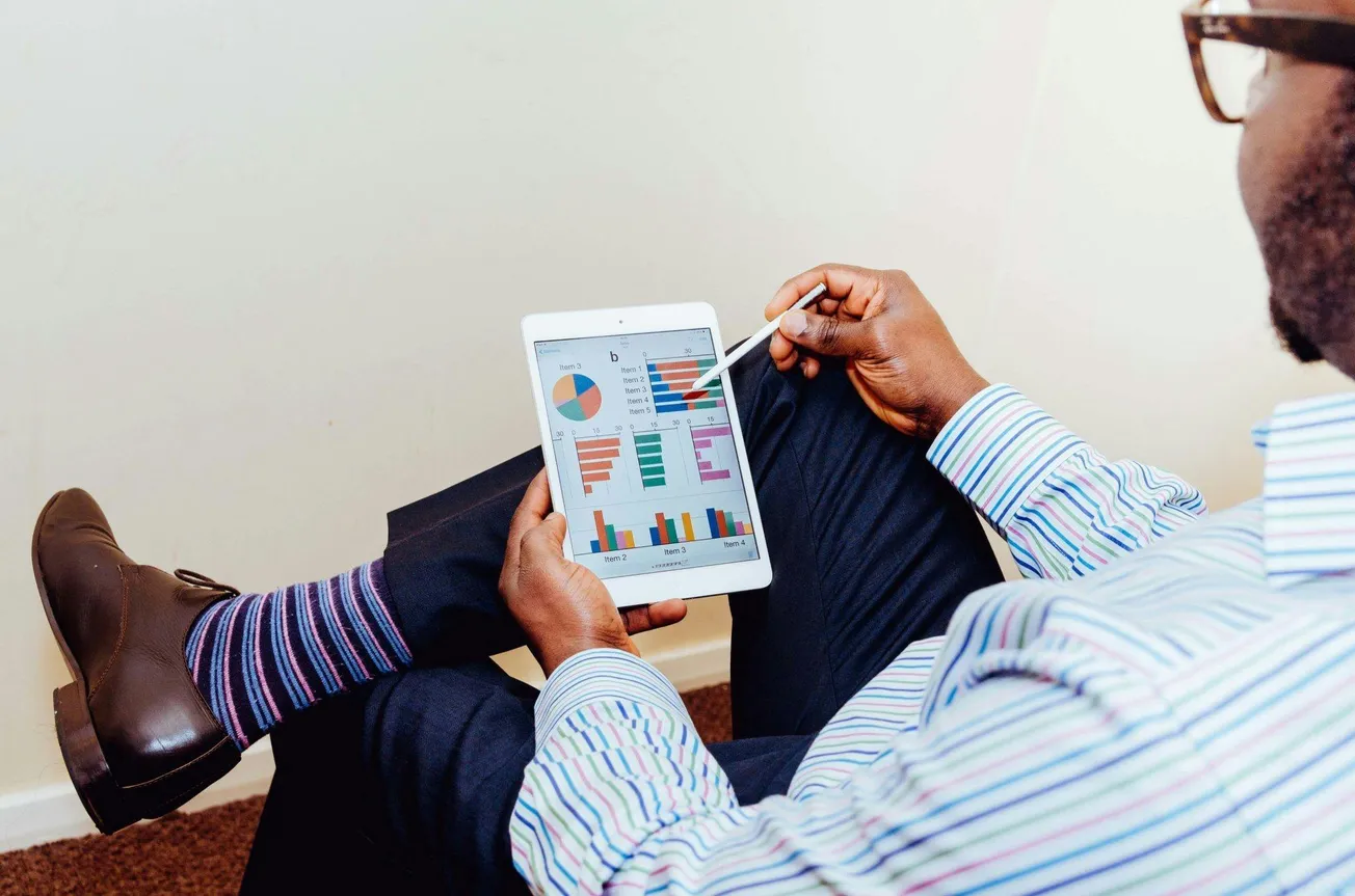 A person in a striped shirt views colorful business graphs and charts on a tablet, using a stylus in a relaxed, seated position with crossed legs.