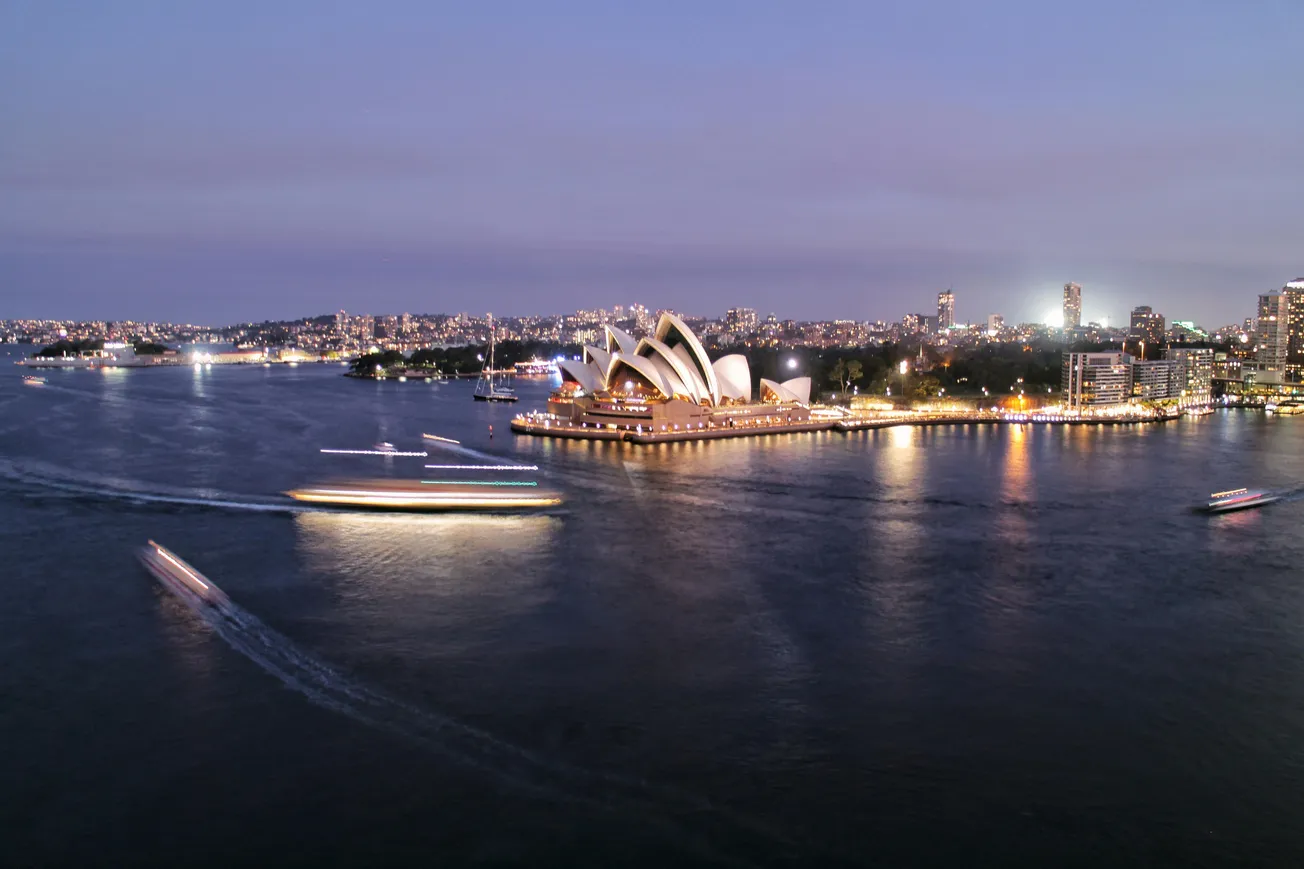 A twilight view of Sydney Harbour, featuring the illuminated Sydney Opera House. Boats with light trails move across the water, highlighting a vibrant cityscape.